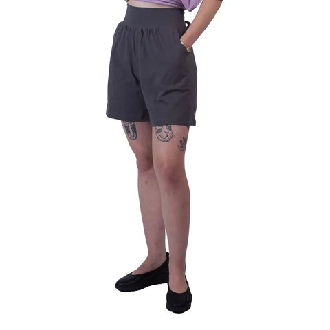 A person wears the Lexi Lounge Shorts by Spiritex | Sustainable Apparel in dark, high-waisted style with a light purple top and black shoes. They stand hands-in-pockets, showing leg tattoos, against a plain white background.