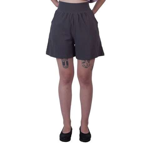 A person models Spiritex | Sustainable Apparels Lexi Lounge Shorts in loose, dark gray organic cotton, paired with black shoes. Tattoos are visible on both thighs against a plain white background, with hands behind their back.