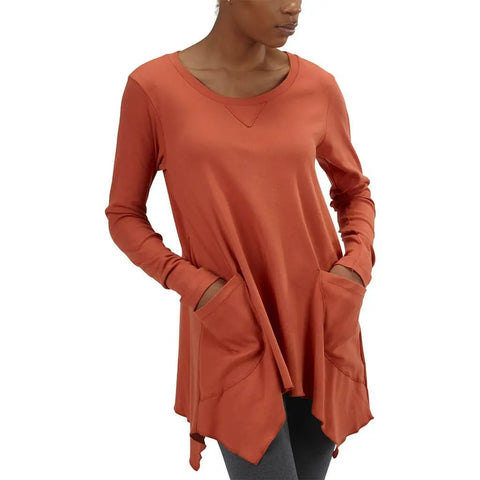 A woman wears the rust-colored Long Sleeve Jenna Hippie Top by Spiritex | Sustainable Apparel, featuring an asymmetrical hem and large front pockets. Made in the USA, the loose tunic drapes over dark leggings as she stands with hands in her pockets.