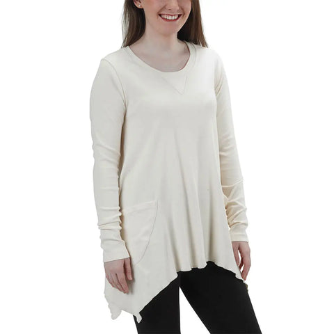 A woman stands smiling against a white background, wearing the off-white Long Sleeve Jenna Hippie Top by Spiritex | Sustainable Apparel. The made-in-USA tunic features an asymmetrical hem, front pocket, and pairs with black pants.