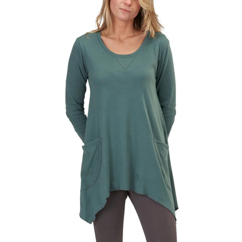 A woman stands with her hands in the pockets of a loose-fitting Long Sleeve Jenna Hippie Top by Spiritex | Sustainable Apparel, featuring two front pockets, long sleeves, and an asymmetrical hem, paired with brown leggings.