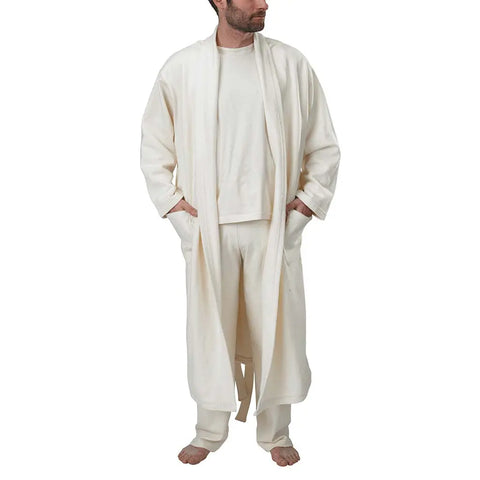 A person stands barefoot, wearing the Spiritex Mid-Length Fleece Robe and loose pants, both in cream organic cotton, with a long-sleeve shirt. The face is turned slightly to the side against a plain white background.