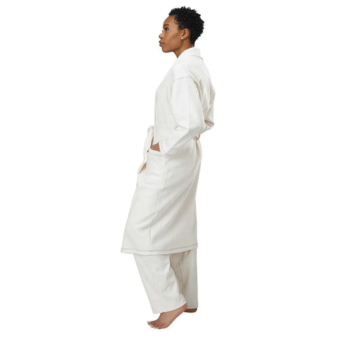 A person stands barefoot in profile, wearing the Spiritex | Sustainable Apparel Mid-Length Fleece Robe in white with matching pants and hands in pockets, set against a plain white background.
