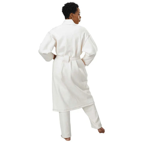 A person stands barefoot with one leg raised, wearing the Spiritex Mid-Length Fleece Robe and matching white pants. Shown from the back, the unisex robe’s loose fit is highlighted against a plain white background.