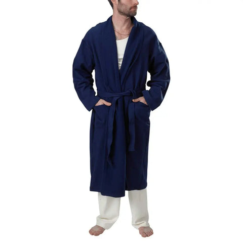 A man in a navy blue Mid-Length Fleece Robe by Spiritex | Sustainable Apparel, featuring a belt, white shirt, and white pants, stands barefoot against a plain white background and looks to his left.