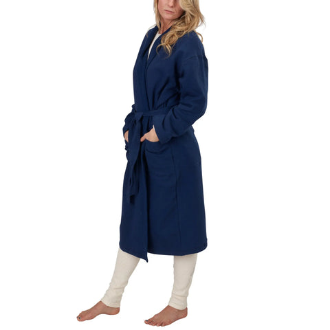 A woman stands barefoot in a navy Mid-Length Fleece Robe by Spiritex | Sustainable Apparel, belted at the waist with her hands in the pockets, cream leggings underneath. She gazes to the side, hair loose and wavy, against a white background.