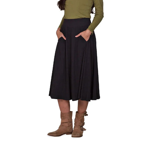 A person wearing a long-sleeve olive top, the Spiritex Gore Skirt by Spiritex | Sustainable Apparel—an organic cotton A-line skirt with pockets—and tan suede ankle boots, stands with hands in the skirt’s pockets against a white background.