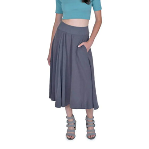 A woman stands against a white background with one hand in her pocket, wearing a teal crop top, gray Spiritex Gore Skirt made of organic cotton with pockets, and strappy gray sandals.