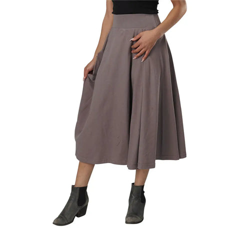 A person models the Gore Skirt by Spiritex | Sustainable Apparel in taupe, paired with a black top and gray ankle boots, standing against a white background with one hand on their hip and the other holding the skirt.