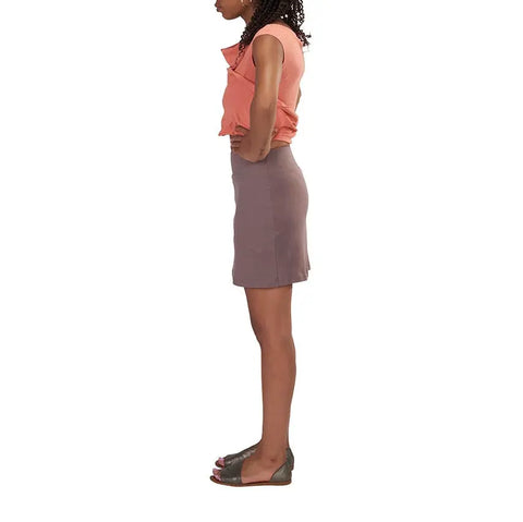 A woman stands in profile against a white background, wearing a coral sleeveless top, the Andy Skirt by Spiritex | Sustainable Apparel in taupe—made from organic cotton—and open-toe flat sandals. Her arms are relaxed at her sides.