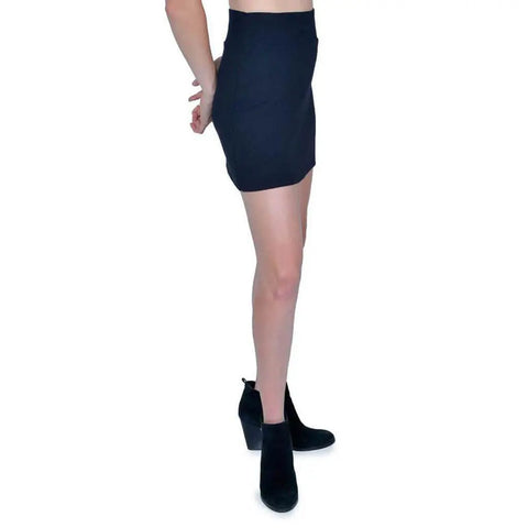 A person stands in profile against a white background, wearing the fitted black Andy Skirt by Spiritex | Sustainable Apparel and chunky-heeled ankle boots—an eco-friendly look. Their arms are bent behind their back.