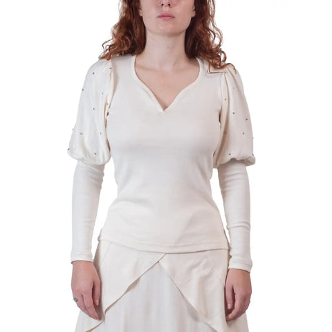 A woman with wavy red hair wears the Victoria Top by Spiritex | Sustainable Apparel, an organic cotton piece with puffed sleeves and silver studs, paired with a matching off-white skirt against a plain white background.