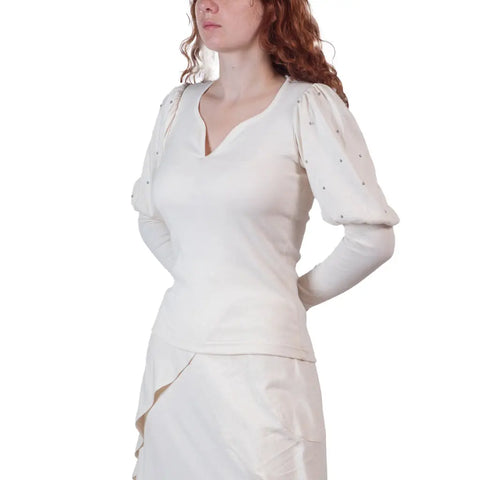 A woman with long curly red hair wears the Spiritex | Sustainable Apparel Victoria Top in organic cotton, with puffed sleeves, and a matching cream skirt. She stands against a white background, her face partially out of frame, hands behind her back.