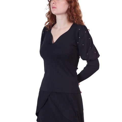 A woman with wavy red hair wears the black Victoria Top from Spiritex | Sustainable Apparel, featuring puffed sleeves and silver studs, paired with a black skirt, standing hands behind her back against a white background.