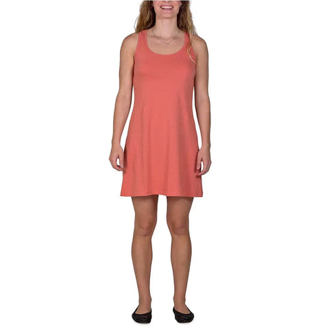 A woman smiles facing forward in a coral Spiritex | Sustainable Apparel Racerback Tank Dress and black flats, ideal for a capsule wardrobe, against a plain white background.