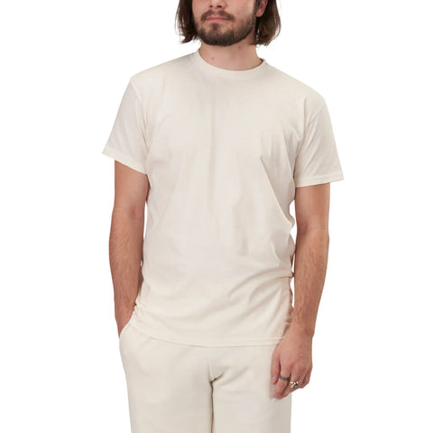A person with shoulder-length brown hair and a beard wears an off-white Short Sleeve Favorite Crewneck Tee (M) by Spiritex | Sustainable Apparel and matching pants, standing with one hand in their pocket against a white background.