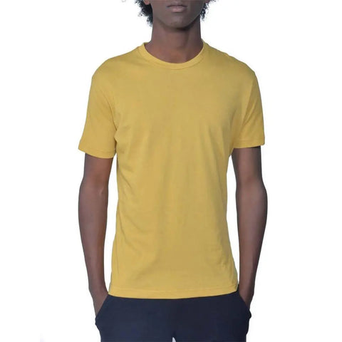 A person with curly hair wears a plain yellow Spiritex | Sustainable Apparel Short Sleeve Favorite Crewneck Tee (M) made from organic cotton and dark pants, standing with hands in pockets against a white background. Face is partially out of frame.