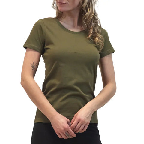 A woman with long wavy hair wears the Spiritex Short Sleeve Favorite Crewneck Tee (W) in olive green and black pants, standing against a white background with hands loosely clasped. A small tattoo is visible on her left upper arm.