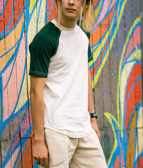 In front of a vibrant graffiti wall, a person with long hair wears the Spiritex | Sustainable Apparel Short Sleeve Baseball Raglan Tee, beige pants, and a black watch, with their face partially out of frame.