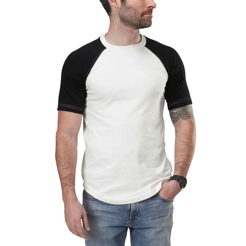 A man with short dark hair and a beard wears a white Spiritex | Sustainable Apparel Short Sleeve Baseball Raglan Tee with black sleeves and blue jeans. He has a watch on his left wrist, a tattoo on his right forearm, and looks to the side with one hand in his pocket.