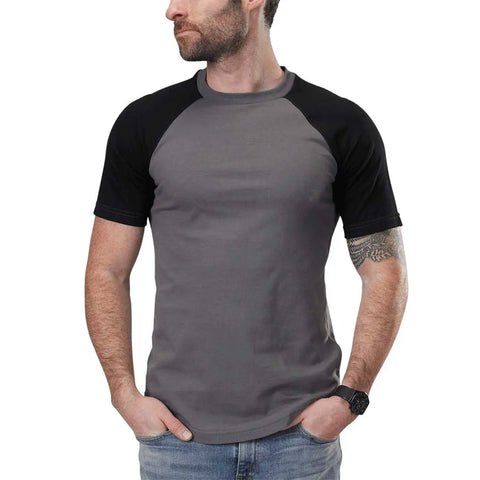 A bearded man stands with hands in pockets, wearing a Spiritex | Sustainable Apparel Short Sleeve Baseball Raglan Tee with black sleeves (100% organic cotton), blue jeans, a left wrist watch, and a right forearm tattoo, looking left.