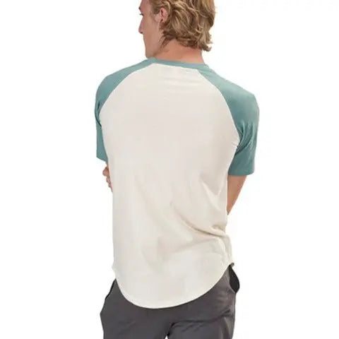A person with blond hair is seen from the back wearing the Spiritex Short Sleeve Baseball Raglan Tee, made from 100% organic cotton with teal sleeves and a cream body, paired with dark shorts, against a plain white background.
