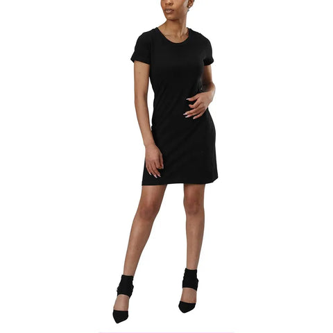A woman poses against a white background in the Spiritex | Sustainable Apparel Short Sleeve Crewneck T-Shirt Dress, pairing it with black heeled shoes. She rests one hand on her hip, the other relaxed, with her head slightly turned to the side.