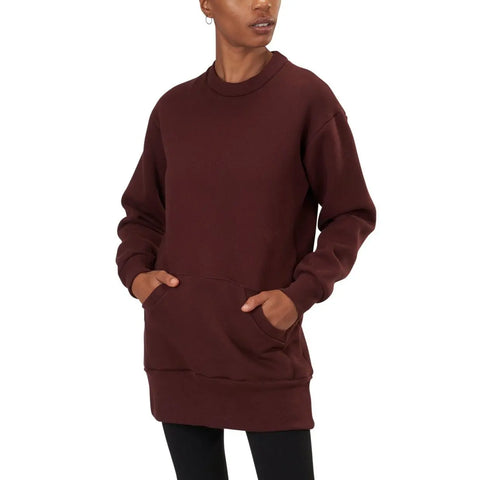 A person wearing the Spiritex | Sustainable Apparel Sweatshirt Tunic, a long, oversized maroon heavyweight fleece with a front pocket, paired with black leggings and standing against a plain white background.