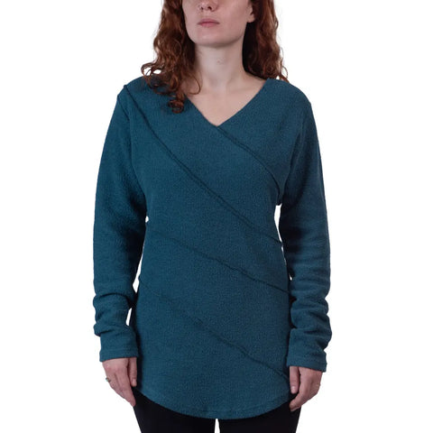 A woman with wavy red hair wears the Spiritex Swirl Pullover, a teal organic cotton V-neck top with long sleeves and diagonal textured lines. She stands against a plain white background.