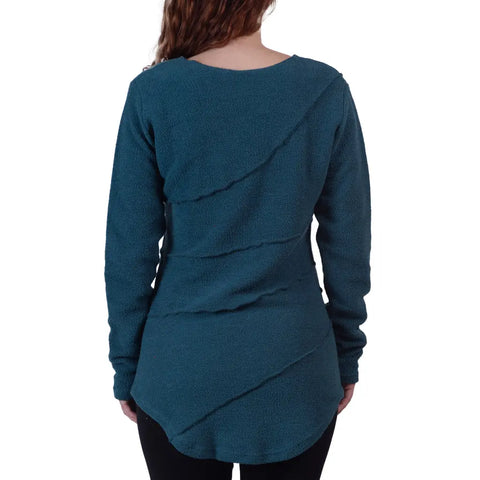 A woman with long brown hair is shown from the back, wearing the Spiritex | Sustainable Apparel Swirl Pullover in teal, a long-sleeve French terry tunic sweater with diagonal seams, paired with black pants.