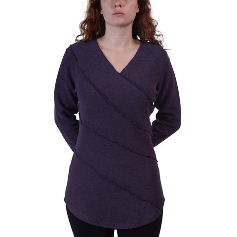 A woman with long, wavy red hair models the Swirl Pullover by Spiritex | Sustainable Apparel—an organic cotton sweater in deep purple with diagonal seam details—standing facing forward, hands behind her back against a white background.