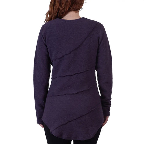 A woman with long, wavy brown hair is seen from behind, wearing the Spiritex Swirl Pullover in dark purple organic cotton with diagonal seams and black leggings.