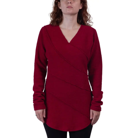 A person with wavy brown hair faces the camera, wearing the Swirl Pullover by Spiritex | Sustainable Apparel—a long-sleeved, deep red organic cotton top with diagonal seam details—standing against a white background.