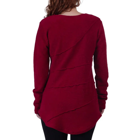A person with long hair is shown from the back wearing the Spiritex Swirl Pullover, a long-sleeve, dark red organic cotton sweater with diagonal seam details, paired with black pants.