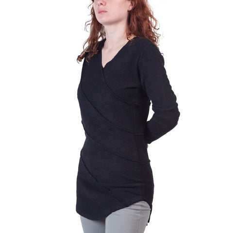 A woman with long curly red hair stands slightly to the side, hands behind her back, wearing the Spiritex Swirl Pullover and light gray pants.