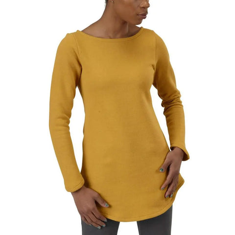 A person wears the Spiritex Maddi Tunic Sweatshirt, a long-sleeve mustard yellow boat neck top, with gray pants—standing with one hand on their hip and the other resting on their thigh.