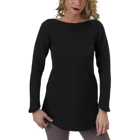 A woman with wavy blonde hair models the Maddi Tunic Sweatshirt by Spiritex | Sustainable Apparel, wearing it in black with mauve leggings, standing against a white background and looking slightly down.