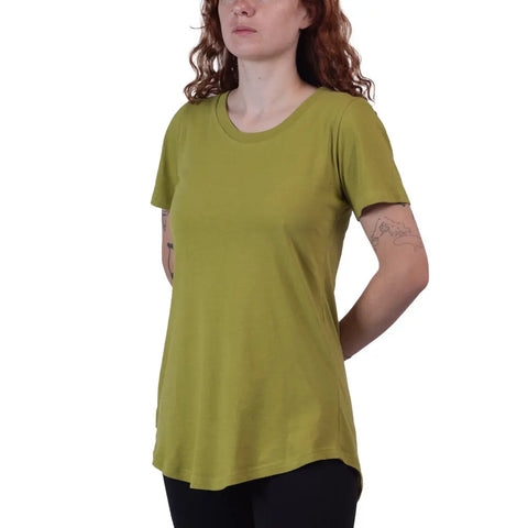 A woman with curly red hair wears the Skyland Short Sleeve Tunic Tee by Spiritex | Sustainable Apparel in light green with black pants, showing tattoos on both arms. She stands relaxed, hands behind her back, and her face is partially visible.
