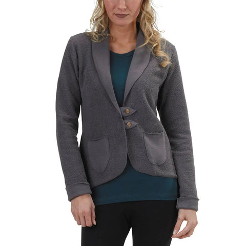 A woman with wavy blonde hair wears the Spiritex Heavyweight Terry Tab Jacket in gray, featuring two front pockets and button closure, over a teal top and black pants. The photo is cropped above her knees.