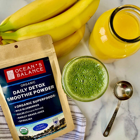 A packet of Oceans Balance Detox Smoothie Powder, a green smoothie, bananas, a mason jar of yellow juice, and a metal tablespoon are arranged on a marble surface.
