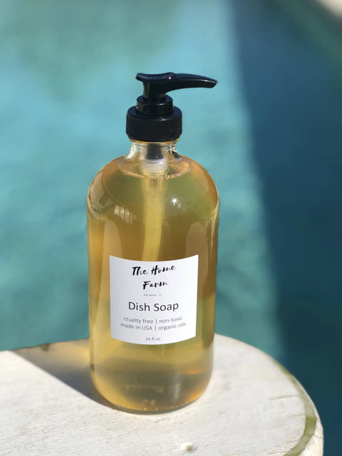 A clear pump bottle labeled The Home Farm Dish Soap rests on a white surface by a pool, highlighting this handcrafted, cruelty-free, non-toxic USA-made dish soap as an eco-friendly cleaner with organic oils.