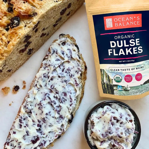 A slice of bread with butter blended with Oceans Balance Organic Dulse Flakes sits beside a bowl of the same spread and a package of Oceans Balance Organic Dulse Flakes, with part of a loaf of bread also shown.