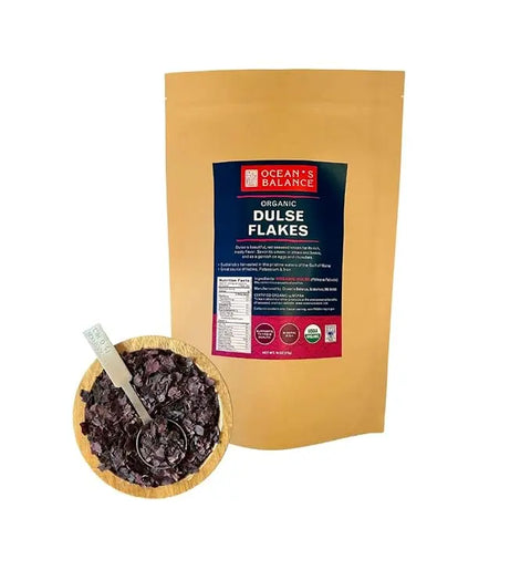 A resealable brown bag labeled Oceans Balance Organic Dulse Flakes stands behind a small wooden bowl filled with dark red seaweed flakes and a metal spoon.