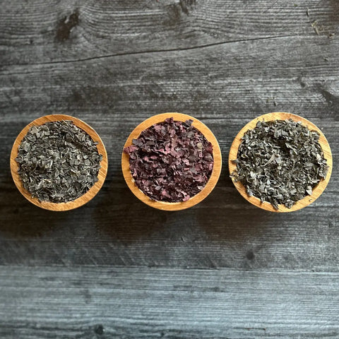 Three small wooden bowls hold assorted dried seaweed, including Oceans Balance Organic Dulse Flakes. Arranged side by side on a dark wood surface, the vibrant USDA Organic selection features dulse and green seaweed flakes.