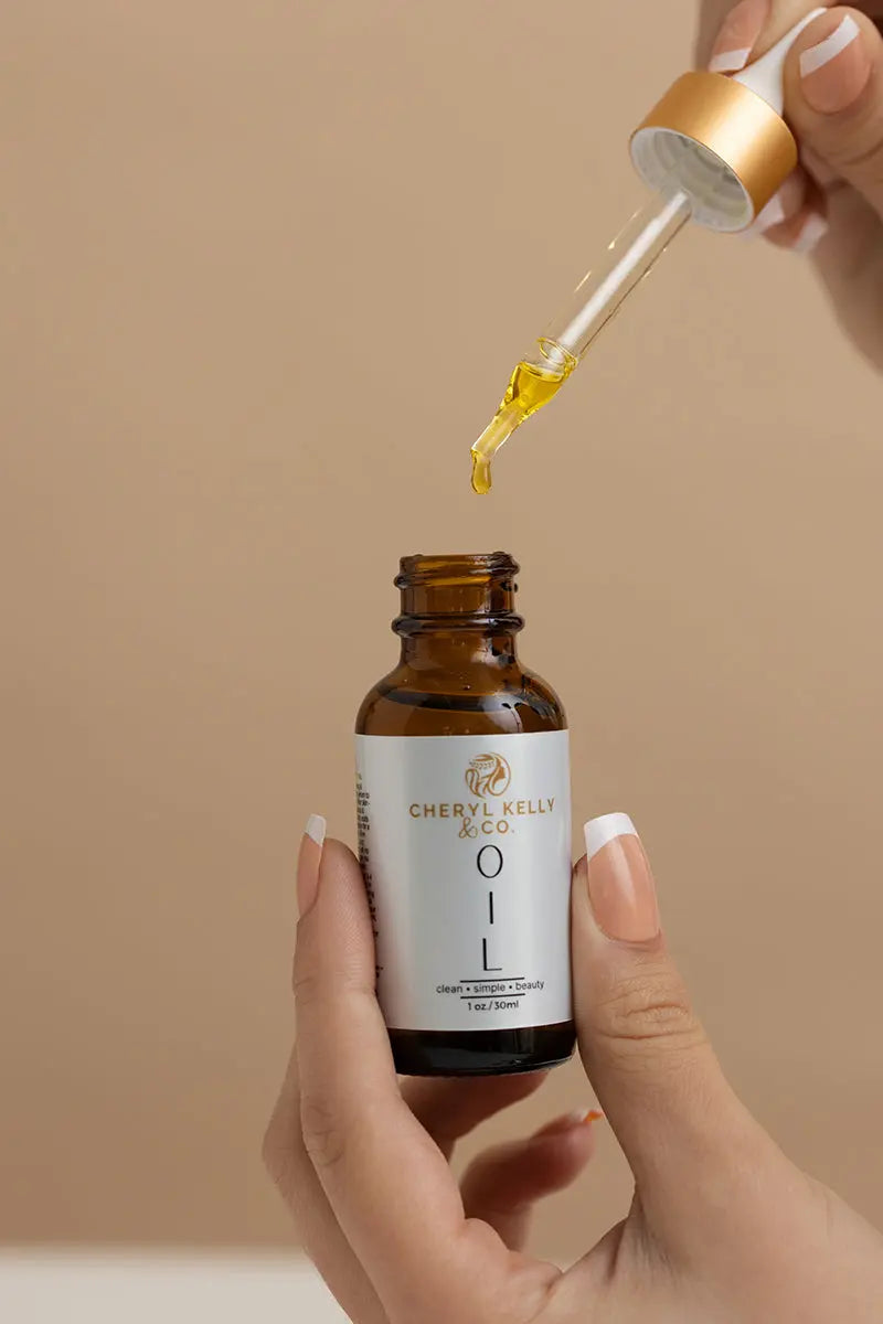 A hand holds an open amber bottle labeled Cheryl Kelly & Co. The Beauty Oil, while another uses a dropper to dispense golden organic oil into it, all set against a beige background.