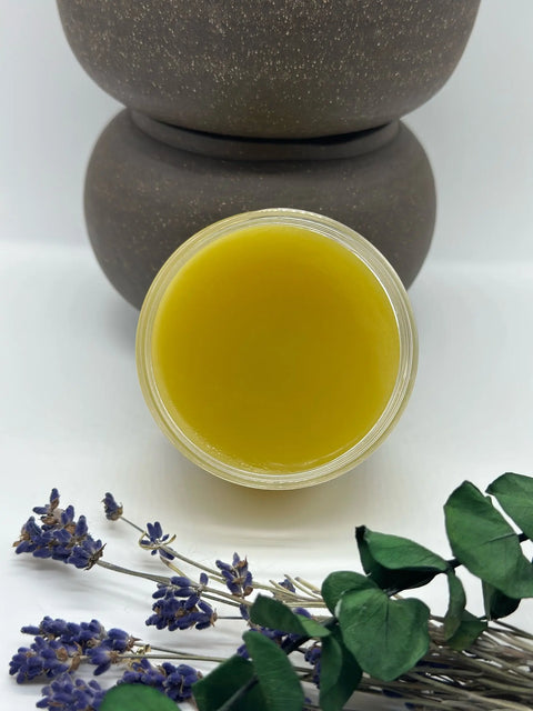 A jar of Five O Clock Co. Organic Nephews Butter is displayed in front of two stacked brown containers, with dried lavender and eucalyptus leaves nearby on a white surface.