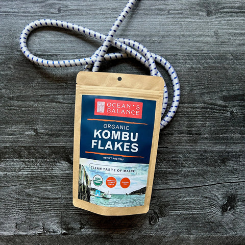A brown resealable pouch of Ocean’s Balance Organic Kombu Flakes sits on a dark wooden surface, with a coiled white and blue rope in the background.