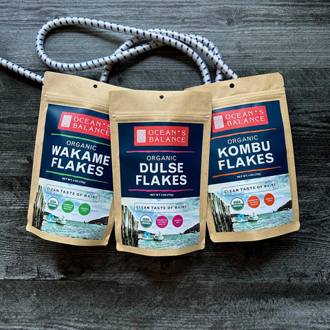 Three brown resealable pouches of Ocean’s Balance seaweed flakes—Wakame, Dulse, and Organic Kombu Flakes—are displayed on a dark wooden surface with a white and blue rope looped behind them.