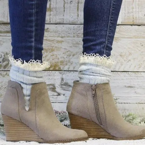 A person wearing Catherine Cole ORGANIC Lace Boot Socks in Carolina Blue, paired with light tan wedge ankle boots and dark skinny jeans, stands on a wooden floor with a rustic wooden backdrop.