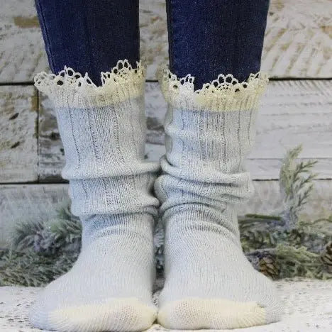 A pair of feet in Catherine Cole ORGANIC Lace Boot Socks – Carolina Blue stands on white, styled with dark blue jeans. Cream trim, rustic wood planks, and greenery set a cozy scene for women’s sock fashion.